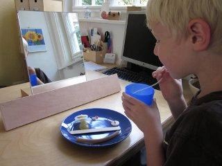 Ein blondes Kind sitzt an einem Schreibtisch, hält einen blauen Becher und betrachtet sich beim Trinken mithilfe eines aufgestellten Spiegels. Auf dem Tisch liegt ein blauer Teller mit Essbesteck und etwas Essen.