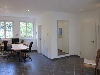 Ein heller, modern eingerichteter Raum mit grauem Fliesenboden, einem großen Spiegel an der Wand, zwei Schreibtischen mit Bürostühlen, Fenstern und weißen Wänden. Ein Computerarbeitsplatz ist erkennbar.