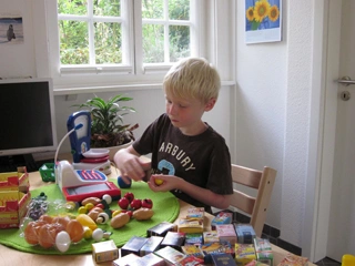 Kind sitzt an einem Tisch und spielt mit Spielzeug-Lebensmitteln und einer Spielkasse, auf dem Tisch liegen viele kleine Verpackungen und Zubehör.