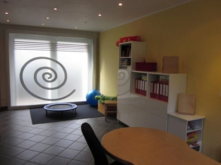 Raum mit großen, satinierten Fenstern mit aufgedruckten Spiralen, einem kleinen Trampolin, Sportball, Regal mit roten Ordnern sowie gelber Wand und Fliesenboden.