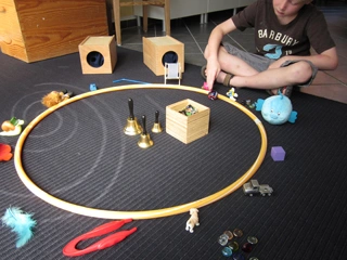 Kind sitzt auf einer schwarzen Matte und spielt mit verschiedenen kleinen Spielsachen, die rund um einen Holzreifen angeordnet sind, darunter Figuren, Glöckchen und Autos.