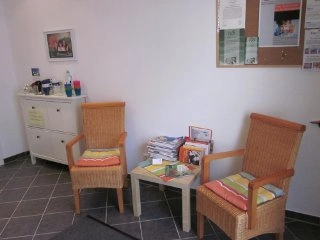 Raum mit zwei geflochtenen Stühlen, bunten Sitzkissen, einem kleinen Tisch mit Zeitschriften und Prospekten sowie einem weißen Sideboard und Infotafel an der Wand.